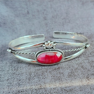 Puede incluir: Un brazalete de plata con una piedra preciosa roja engastada en un bisel ovalado de plata. El brazalete tiene un diseño de alambre trenzado y un detalle decorativo en forma de corona.