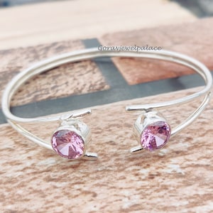 Puede incluir: Un brazalete de plata con dos piedras preciosas rosas. El brazalete está abierto y tiene un diseño sencillo.