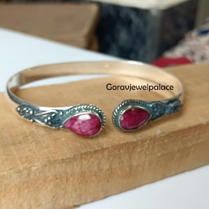 以下が含まれることがあります： シルバーカラーのカフブレスレット。涙型の深紅色の宝石が特徴です。宝石の周りには、装飾的な濃い青緑色のアクセントがあります。「Goravjewelpalace」の文字が木製の表面に表示されています。