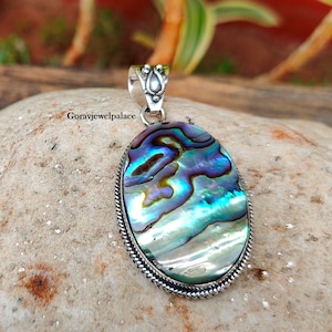 May include: A silver pendant featuring a large oval abalone shell with iridescent blue, green, and purple colors. The shell is set in a silver frame with a detailed border.