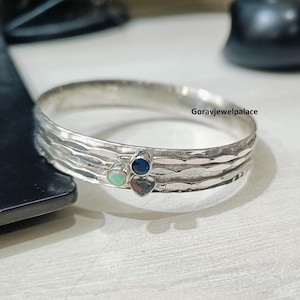 May include: A silver bangle bracelet with a textured surface. The bracelet features three small gemstones: an opal, a dark blue sapphire, and a heart-shaped stone. The bracelet is displayed on a light-colored surface.