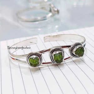 May include: Silver bracelet with three green gemstone accents. The bracelet is open and has a simple design.