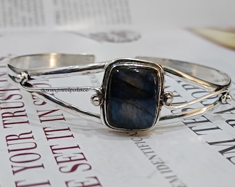 Puede incluir: Pulsera de plata con una piedra preciosa de labradorita azul oscuro. La pulsera tiene un diseño sencillo con dos bandas que se cruzan.