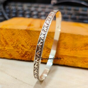 May include: A silver bangle bracelet with a repeating pattern of small fish designs. The bracelet is on a wooden surface.