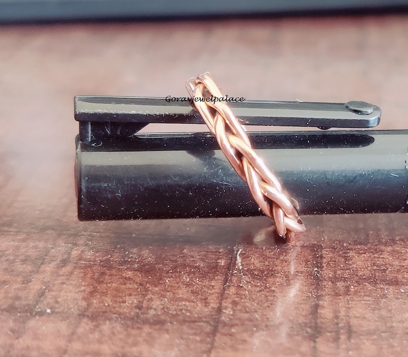 May include: A braided copper ring, with a rose gold finish, is shown on a black surface. The ring is made of multiple strands of copper wire that are woven together to create a braided design.