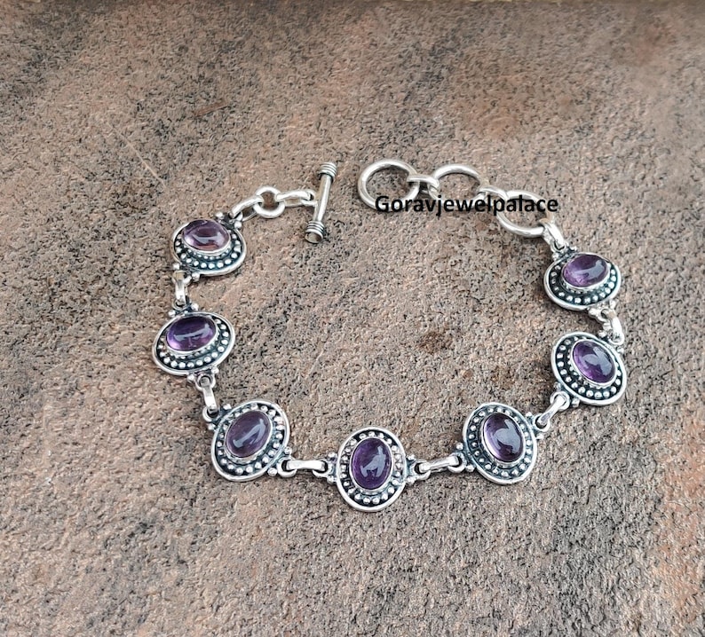 Peut inclure: Bracelet en argent orn&eacute; de pierres d'am&eacute;thyste ovales. Chaque am&eacute;thyste est sertie dans une bordure perl&eacute;e. Le bracelet est dot&eacute; d'un fermoir &agrave; bascule. Le texte "Goravjewelpalace" est visible.
