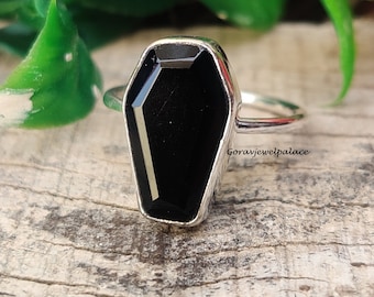 May include: A silver ring featuring a black coffin-shaped gemstone. The ring is displayed on a wooden surface.