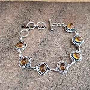 Peut inclure: Bracelet argent&eacute; orn&eacute; de pierres pr&eacute;cieuses ovales de couleur ambre. Le bracelet est dot&eacute; d'un fermoir &agrave; bascule et est compos&eacute; de maillons interconnect&eacute;s. Chaque pierre est sertie dans un bord d&eacute;coratif et perl&eacute;. Sur une surface brune textur&eacute;e.
