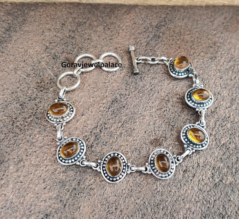 Peut inclure: Bracelet argent&eacute; orn&eacute; de pierres pr&eacute;cieuses ovales de couleur ambre. Le bracelet est dot&eacute; d'un fermoir &agrave; bascule et est compos&eacute; de maillons interconnect&eacute;s. Chaque pierre est sertie dans un bord d&eacute;coratif et perl&eacute;. Sur une surface brune textur&eacute;e.