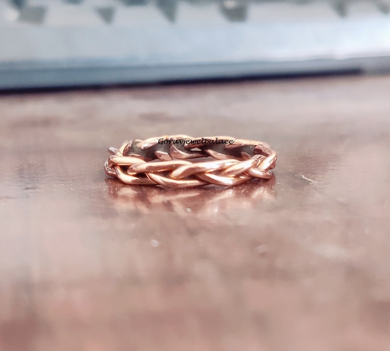 May include: A rose gold braided ring with a woven design. The ring is on a wooden surface.