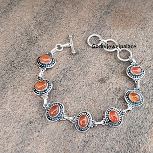 Peut inclure: Un bracelet en argent orn&eacute; de pierres pr&eacute;cieuses ovales, rouge-orang&eacute;, serties dans un motif de lunette d&eacute;coratif. Le bracelet est dot&eacute; d'un fermoir &agrave; bascule et est pr&eacute;sent&eacute; sur une surface textur&eacute;e de tons neutres. Un accessoire &eacute;l&eacute;gant.