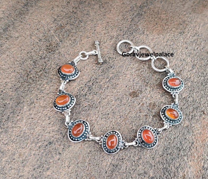 Peut inclure: Un bracelet en argent orn&eacute; de pierres pr&eacute;cieuses ovales, rouge-orang&eacute;, serties dans un motif de lunette d&eacute;coratif. Le bracelet est dot&eacute; d'un fermoir &agrave; bascule et est pr&eacute;sent&eacute; sur une surface textur&eacute;e de tons neutres. Un accessoire &eacute;l&eacute;gant.