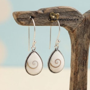 May include: A pair of drop earrings featuring teardrop-shaped shiva eye shells with a natural spiral pattern. Each shell is set in a silver-toned metal frame with a twisted rope border, suspended from long, curved silver-toned metal ear wires.
