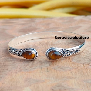 May include: Silver bangle bracelet with two tiger's eye stones. The bracelet has a delicate design with small beads and a twisted wire detail. "Goravijewelpalace" is written on the bracelet.