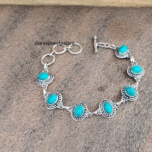 Peut inclure: Un bracelet en argent orn&eacute; de pierres ovales turquoise serties dans un design de lunette d&eacute;taill&eacute;. Le bracelet a un fermoir &agrave; bascule et est pr&eacute;sent&eacute; sur une surface marron textur&eacute;e. Le texte "Goravjewelpalace" est visible.