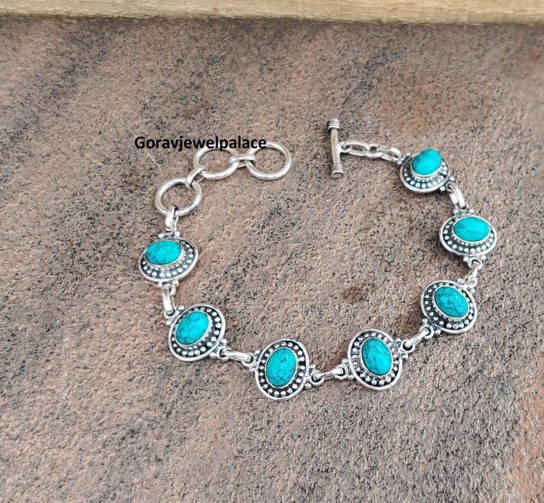 Peut inclure: Un bracelet en argent orn&eacute; de pierres ovales turquoise serties dans un design de lunette d&eacute;taill&eacute;. Le bracelet a un fermoir &agrave; bascule et est pr&eacute;sent&eacute; sur une surface marron textur&eacute;e. Le texte "Goravjewelpalace" est visible.