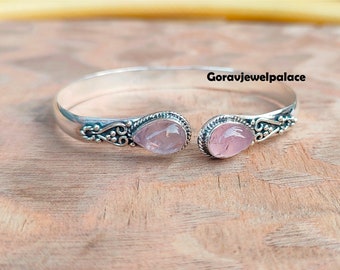 May include: Silver bangle bracelet featuring two pink rose quartz stones. The bracelet has an open design and intricate detailing.