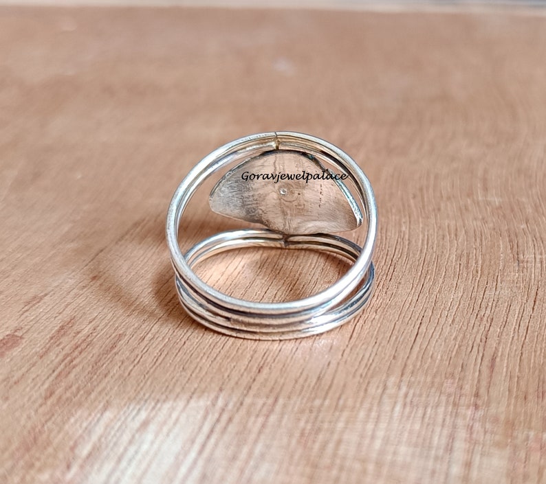 May include: A silver ring with a layered band design. The ring features a central, oval-shaped stone with a textured surface. The brand name "Goravjewelpalace" is visible on the stone. The ring is set against a wooden background.