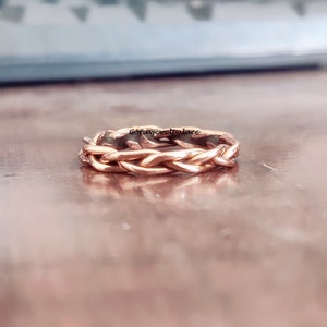May include: A rose gold braided ring with a woven design. The ring is on a wooden surface.
