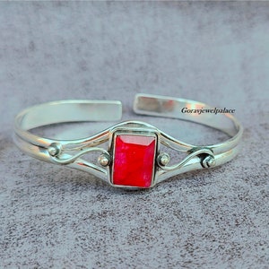 Puede incluir: Un brazalete de plata con una piedra preciosa roja engastada en un marco de plata. El brazalete tiene un diseño delicado y en espiral.