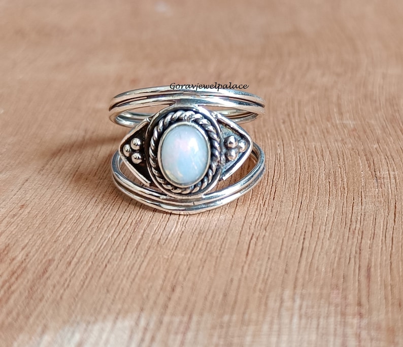 May include: A silver ring with an oval, opalescent stone at the center. The stone is surrounded by a twisted rope design and decorative accents. The ring has a layered band design and is set against a wooden surface.