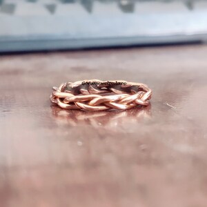 May include: A rose gold braided ring with a woven design. The ring is on a brown surface.