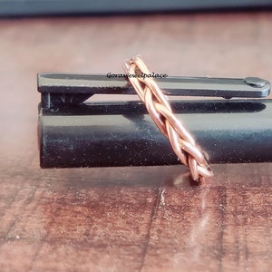 May include: A braided copper ring, with a rose gold finish, is shown on a black surface. The ring is made of multiple strands of copper wire that are woven together to create a braided design.