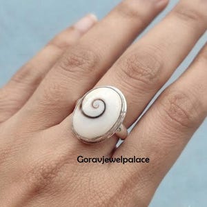 Könnte beinhalten: Ein Silberring mit einer ovalen Shiva-Auge-Muschel, die ein natürliches braunes Spiralmuster aufweist und in ein poliertes Metallband gefasst ist. Der Text Goravjewelpalace ist auf dem Bild sichtbar. Das Design betont die organische Spirale.