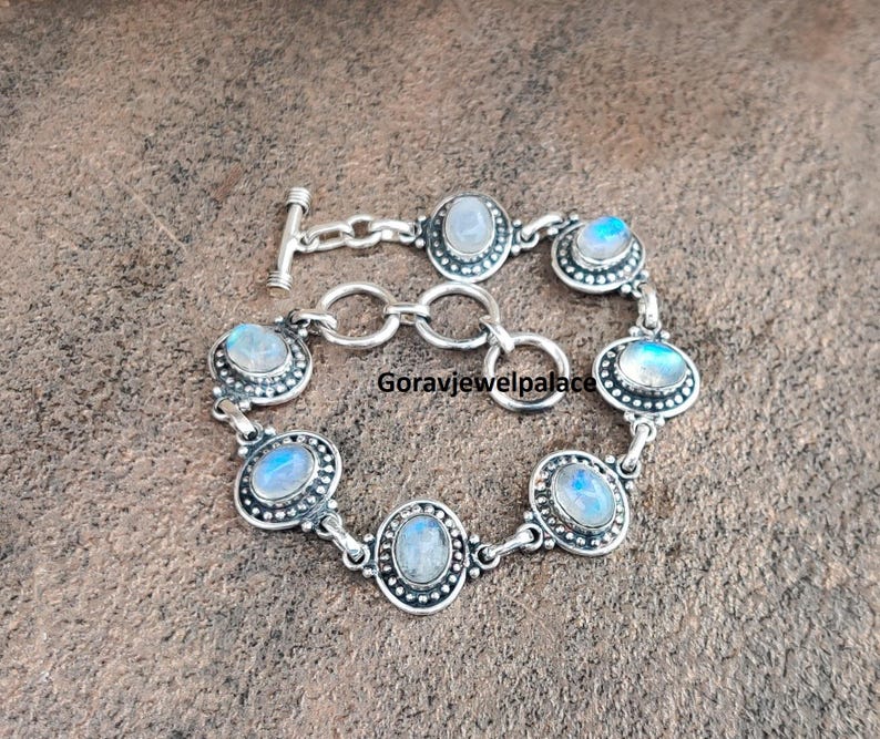Peut inclure: Un bracelet en argent orn&eacute; de cabochons de pierres de lune ovales et ronds. Le bracelet est dot&eacute; d'un fermoir &agrave; bascule et est compos&eacute; de maillons interconnect&eacute;s. Les pierres de lune ont une lueur iris&eacute;e bleue et blanche.