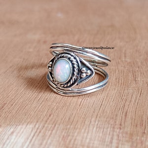 May include: A silver ring with an oval opal gemstone at the center. The opal has a pearly white color with hints of pink and orange. The ring has a detailed, textured design around the gemstone and on the band. The ring is displayed on a wooden surface.