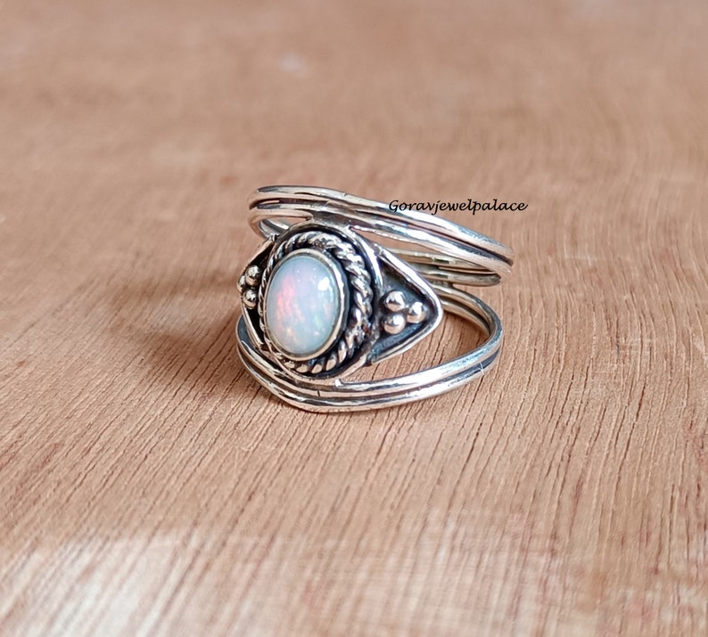 May include: A silver ring with an oval opal gemstone at the center. The opal has a pearly white color with hints of pink and orange. The ring has a detailed, textured design around the gemstone and on the band. The ring is displayed on a wooden surface.