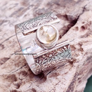 May include: A silver ring with a floral design and a clear gemstone in the center. The ring is wide and has a unique design.