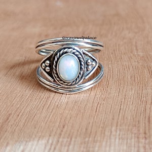 May include: A silver ring with an oval, opalescent stone at the center. The stone is surrounded by a twisted rope design and decorative accents. The ring has a layered band design and is set against a wooden surface.