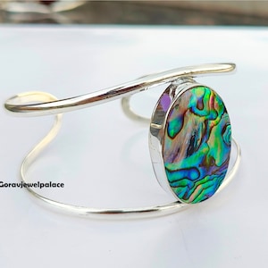 Puede incluir: Un brazalete de plata con una gran incrustación ovalada de concha de abulón. La concha tiene colores iridiscentes de verde, azul, morado y marrón.