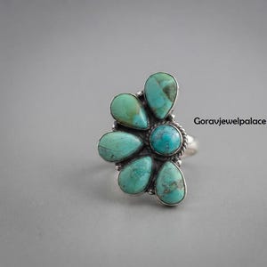 Peut inclure: Une bague en argent avec une pierre turquoise ronde centrale entourée de cinq pierres turquoise en forme de goutte disposées en éventail. L'anneau en métal présente une texture torsadée. Le texte Goravjewelpalace est visible à droite.