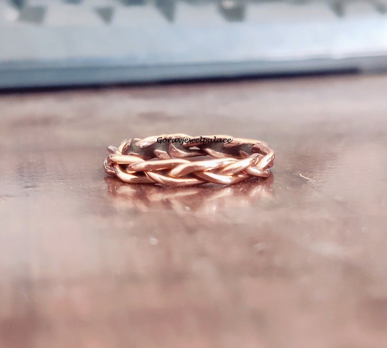 May include: A rose gold braided ring with a woven design. The ring is made of metal and has a shiny finish.
