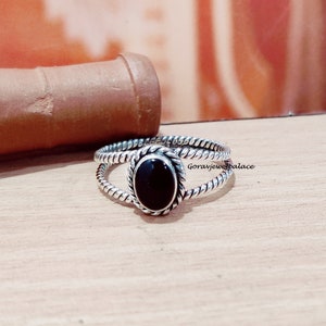 May include: A silver ring with a black oval gemstone set in a twisted band. The ring is on a wooden surface.