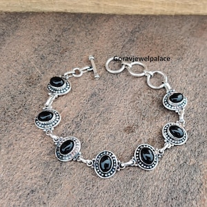 Peut inclure: Bracelet en argent orn&eacute; de pierres noires ovales. Le bracelet est dot&eacute; d'un fermoir &agrave; bascule et est compos&eacute; de maillons interconnect&eacute;s. Chaque pierre est sertie dans un cadre d&eacute;coratif perl&eacute;. Le bracelet est pr&eacute;sent&eacute; sur une surface brune textur&eacute;e.