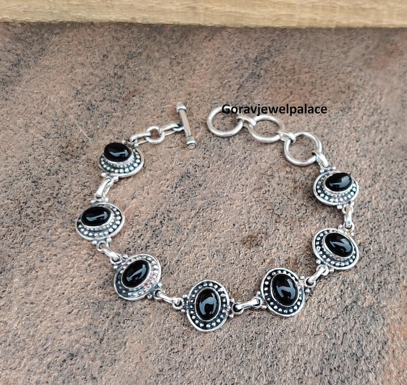 Peut inclure: Bracelet en argent orn&eacute; de pierres noires ovales. Le bracelet est dot&eacute; d'un fermoir &agrave; bascule et est compos&eacute; de maillons interconnect&eacute;s. Chaque pierre est sertie dans un cadre d&eacute;coratif perl&eacute;. Le bracelet est pr&eacute;sent&eacute; sur une surface brune textur&eacute;e.