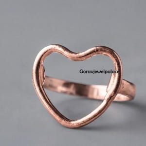 May include: A copper-toned ring featuring an open heart-shaped design. The metal surface has a slightly hammered, textured appearance. The text Goravjewelpalace is visible on the right side of the heart frame, which is centred on the band.