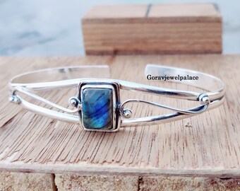 Puede incluir: Pulsera de brazalete de plata con una piedra preciosa de labradorita azul. La pulsera tiene un diseño delicado y curvo con una sola piedra preciosa en el centro.