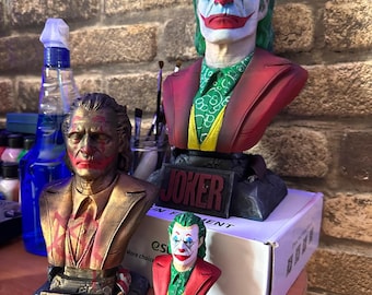 Könnte beinhalten: Drei Joker-Büsten, eine große, eine mittlere und eine kleine, alle in Rot, Grün und Gelb bemalt. Die Büsten stehen auf einer Holzoberfläche mit einer weißen Box dahinter. Die größte Büste hat das Wort "JOKER" auf dem Sockel.