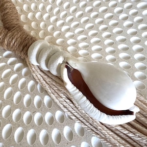 May include: A decorative seashell in natural brown and white tones. The shell is attached to a loop made of brown rope, with a white, textured base and a bundle of beige rope strands.