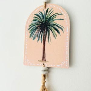Op de afbeelding: Een roze en witte palmboom wandhanger met een kwast van natuurlijke vezels. De palmboom is geschilderd in aquarelstijl.