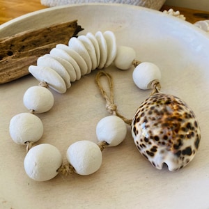 May include: A decorative beaded garland featuring white wooden beads, a section of flat, round white beads, and a speckled seashell. The garland is strung on natural twine and rests in a white wooden bowl, evoking a coastal theme.