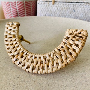 May include: A natural woven rattan cabinet knob with a brass screw. The knob is shaped like a half-circle and has a woven pattern.