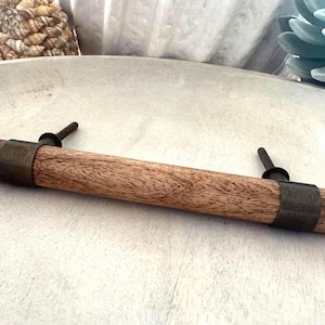 May include: A cylindrical wooden cabinet handle featuring a natural brown wood grain finish, accented by two dark metallic bands and two matching threaded metal bolts extending from the back for mounting purposes.
