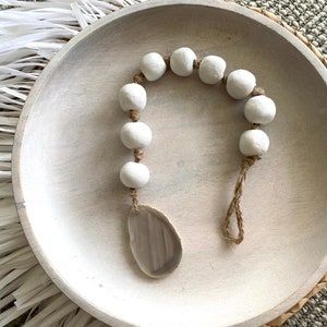 May include: A white ceramic bead garland with a natural twine string and a large, light brown agate slice pendant. The garland is displayed in a white wooden bowl.