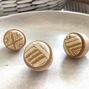 Puede incluir: Tres pomos redondos de madera con un patrón tejido en beige y blanco. Los pomos están sobre una superficie marrón claro.