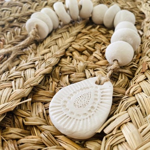 Pode incluir: Uma guirlanda de cerâmica branca com um pingente decorativo em forma de lágrima. A guirlanda é feita de contas redondas e é enfiada em um barbante natural. A guirlanda é exibida em uma superfície de palha tecida.